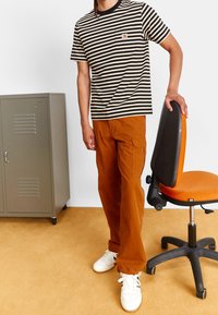 T-shirt rayé noir et beige avec poche, associé à un pantalon cargo orange. Baskets blanches. Casier gris et chaise de bureau orange en arrière-plan.