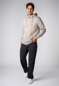 Lichtgrijze rits hoodie met koordjes en zakken aan de voorkant, gecombineerd met losse zwarte broeken en witte sneakers. Eenvoudig en neutraal ontwerp.