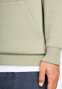 Primo piano di una persona che indossa una felpa verde chiaro con polsini e fondo a costine, una maglietta bianca sotto e pantaloni blu scuro con texture.