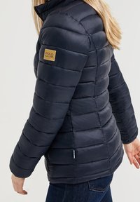 Marinblå pufferjacka med hög krage, vadderad textur och ett litet guld "POLO CLUB"-märke på vänster axel. Mjuk, lätt material.