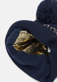 Jūras armijas beanie cepure ar pom-pomu; iekšējā daļa ar mirdzošu zelta sietu; mīksta adīta tekstūra; ribota apmale.