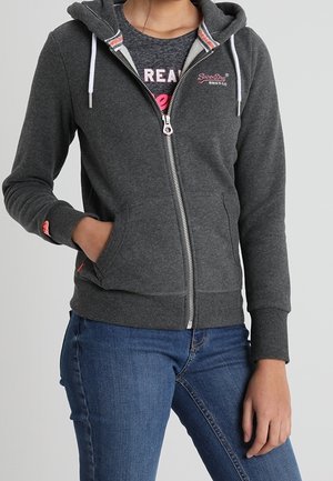 Persoon draagt een donkergrijze zip-up hoodie met witte trekkoorden over een grafisch T-shirt en blauwe spijkerbroek, met één hand in de zak, staand tegen een lichte achtergrond.