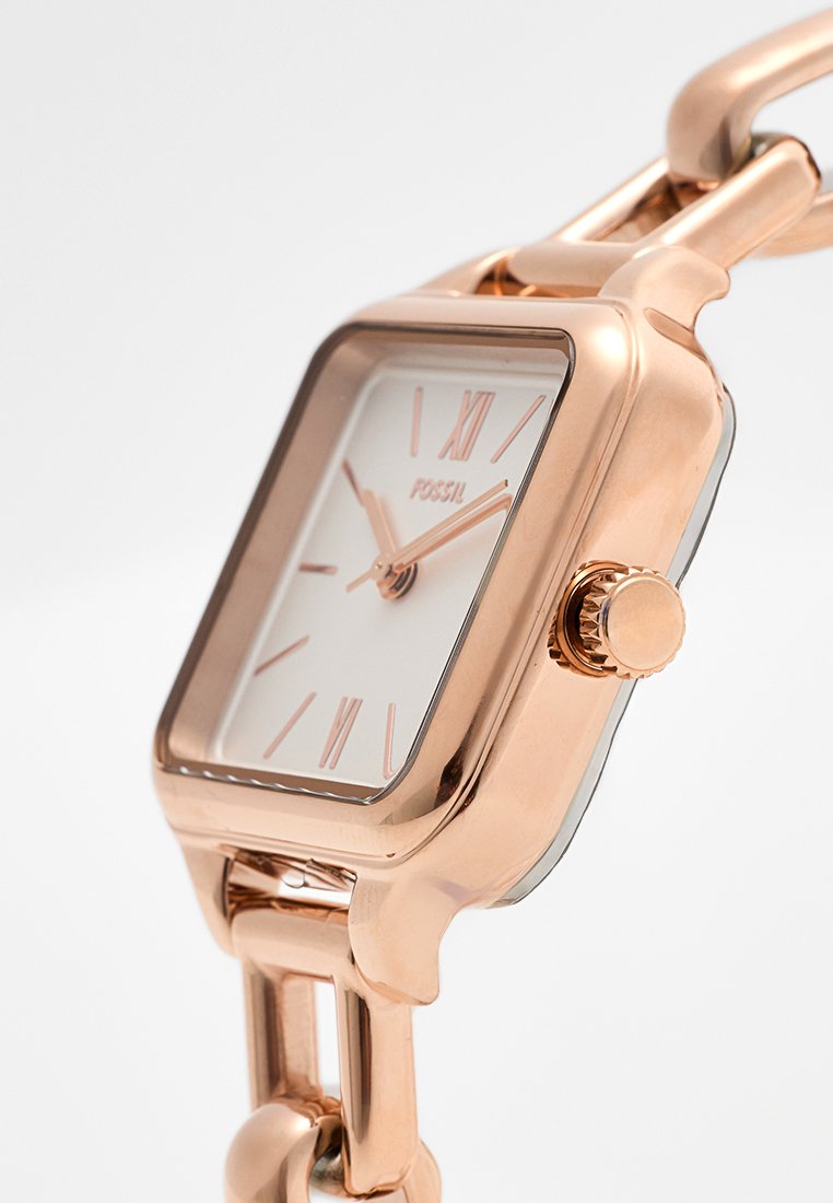 Orologio in metallo rosa con quadrante quadrato, quadrante bianco, numeri romani, lancette sottili e una corona prominente. Presenta un bracciale a maglie grosse.