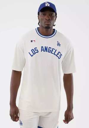 Mann trägt weißes Los Angeles Baseball-Trikot und passende Kappe, steht mit neutralem Gesichtsausdruck vor einfachem Hintergrund.