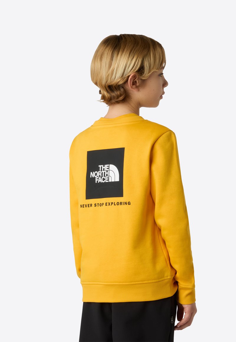 Gul hettegenser laget av mykt stoff, med en stor svart kvadratisk logo på baksiden med teksten "NEVER STOP EXPLORING" under.