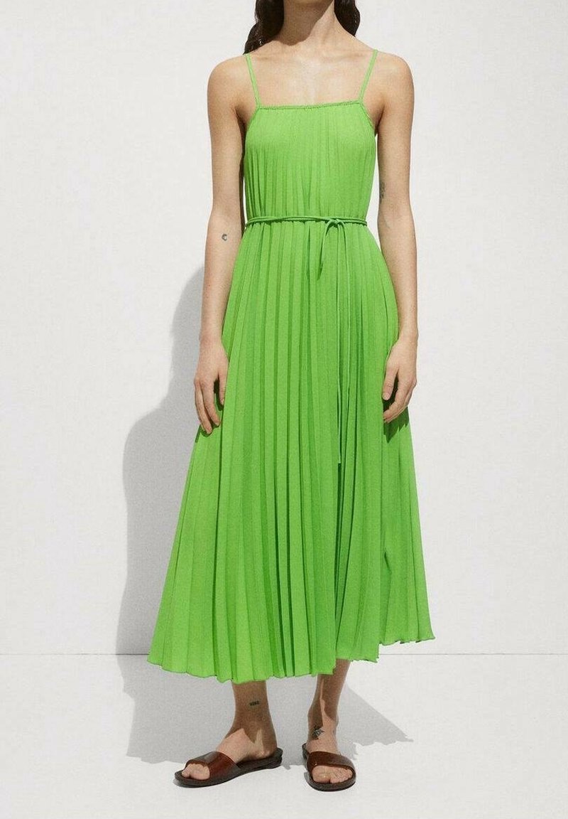Femme portant une robe midi plissée sans manches de couleur vert citron avec une ceinture fine, associée à des sandales plates marron.