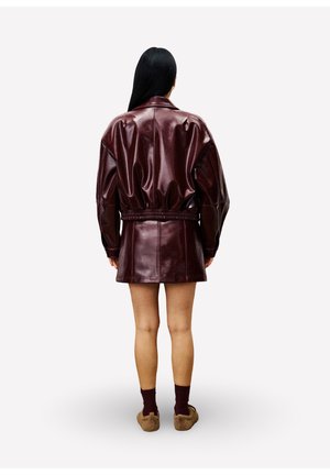 Une personne avec de longs cheveux noirs portant une veste en cuir bordeaux foncé brillante et une jupe assortie, des chaussettes bordeaux et des chaussures en peluche marron, debout.
