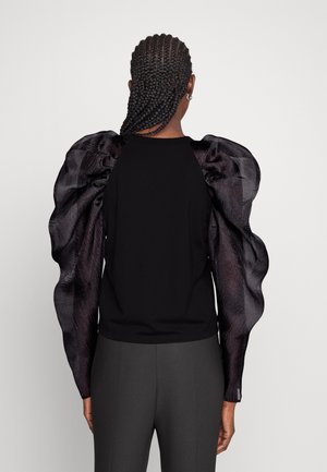 Persona con cabello trenzado que lleva una blusa negra con mangas abullonadas, translúcidas y voluminosas, y pantalones oscuros, vista desde atrás.