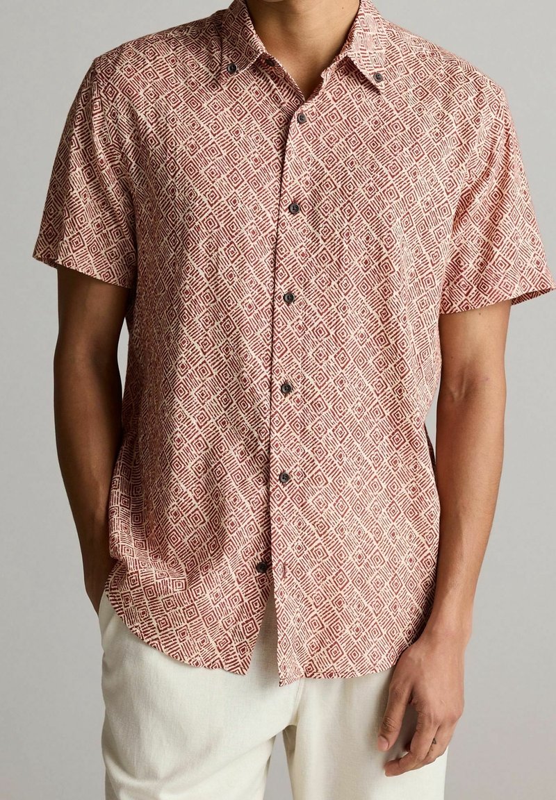 Chemise à manches courtes en bordeaux avec un motif en losanges. Fabriquée en tissu léger, elle comporte des boutons noirs et une coupe décontractée.