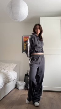 Junge Frau steht in einem Schlafzimmer, trägt einen dunkelgrauen Trainingsanzug mit weißen Streifen und weiße Hausschuhe, neben einem Bett und einem gerahmten Poster.