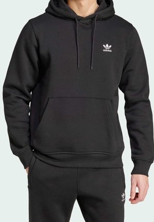 Mężczyzna noszący czarną bluzę z kapturem Adidas i dopasowane czarne spodnie dresowe, przód z widocznym logo na obu częściach odzieży.