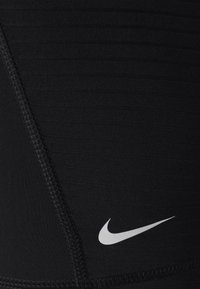 Musta urheiluvaatekangas, jossa on teksturoitu, uurrettu kuvio. Siinä on valkoinen Nike-swoosh-logo. Saumojen varrella on näkyvissä ompelu yksityiskohtia.