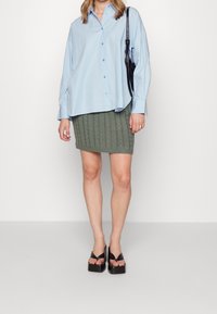 Camisa de botones azul claro, falda de punto verde oliva con patrón de trenzas y sandalias negras de plataforma. Conjunto casual con un corte moderno.