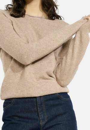 Persoon die een beige, lange mouwen gebreide trui over het bovenlichaam aantrekt, draagt donkere blauwe spijkerbroek, tegen een effen witte achtergrond.