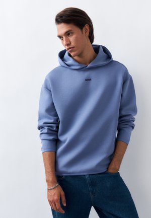 Junger Mann mit mittellangen dunklen Haaren, der einen blauen Hoodie und Jeans trägt und mit einer Hand in der Tasche vor einer weißen Wand steht.