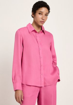 Person with short curly hair wearing a loose-fitting long-sleeve pink button-up shirt and matching pink pants against a plain background.