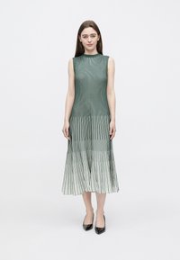 Vestido fluido sem mangas com pregas verticais, que vai do verde escuro no topo ao verde mais claro na bainha, combinado com saltos pretos.