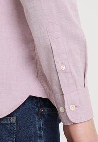 Hellrosa langärmeliges Hemd mit geknöpftem Manschettenverschluss, kontrastierenden Nähten und abgerundetem Saum, kombiniert mit dunkler Jeanshose.