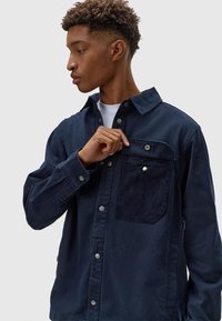 Giacca camicia in corduroy blu navy con chiusura a bottoni, bottoni a scatto, colletto a risvolto e taschino sul petto con una toppa in corduroy.