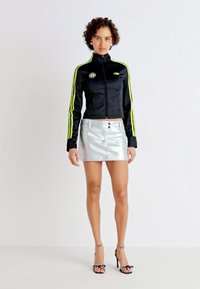 Jachetă neagră scurtă cu dungi verzi neon, asociată cu o fustă mini metalică argintie cu butoane și pantofi cu toc înalt negri.