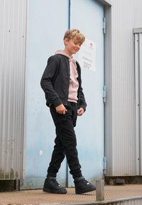 Veste bomber noire sur un hoodie rose, associée à un pantalon cargo noir et des baskets montantes noires. Se tient sur des marches près d'une porte bleue.