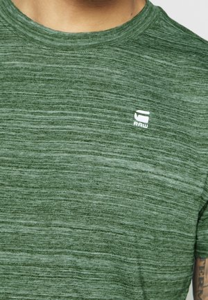 Zielona heatherowana koszula z krótkim rękawem z białym logo "RAW" na piersi, noszona przez osobę, widoczne częściowe tatuaże na ramieniu.