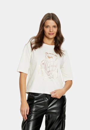 T-shirt bianca corta con grafica di un orso e design a forma di cuore in colori metallici. Maniche corte e vestibilità rilassata, abbinata a pantaloni neri.