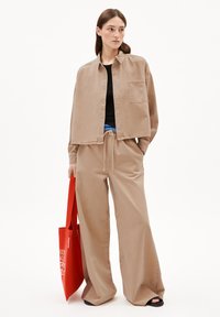 Beige denim jack met een zak, gecombineerd met bijpassende wijde broek, zwart topje en een rode tote bag. Platte sandalen maken de look af.