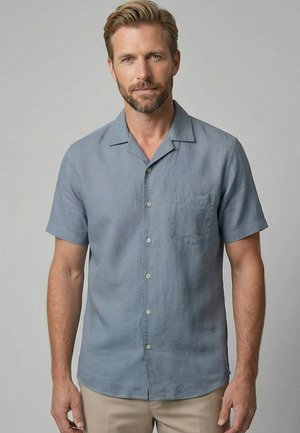 Man met baard die een lichtblauw overhemd met korte mouwen en een beige broek draagt, staand tegen een effen achtergrond.