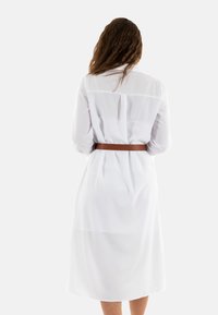 Robe blanche légère avec une taille ceinturée, des manches longues et une longueur mi-mollet. Présente un col classique et des détails en boutons au dos.