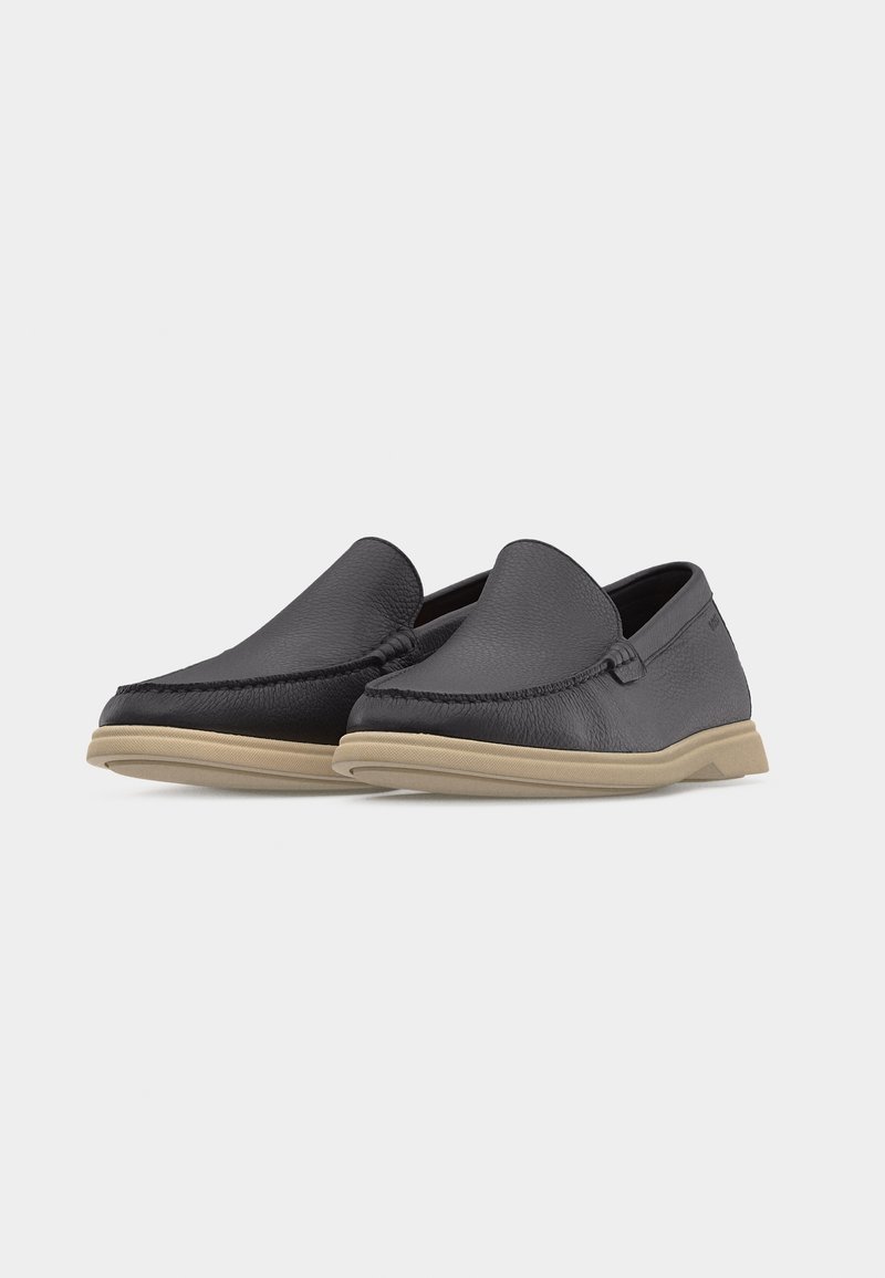 Paire de mocassins en cuir noir sans lacets avec semelles en caoutchouc beiges, bouts arrondis et détails de piqûres subtils sur fond blanc.