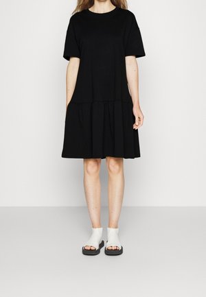 Schwarzes kurzärmeliges Kleid aus Baumwolle mit einem gerafften Stufensaum am unteren Ende. Kombiniert mit weiß-schwarzem, modernem Schuhwerk im minimalistischen Design.