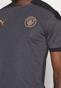 Gray sports jersey with black accents, featuring the Manchester City logo in copper. Made of smooth, breathable fabric with textured details on sleeves.