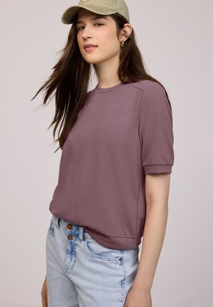 Jeune femme aux longs cheveux bruns portant un t-shirt mauve à manches courtes, un jean bleu clair, une casquette beige et des boucles d'oreilles argentées en forme de créoles.