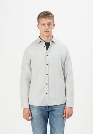 Camisa de botones de color gris claro con cuello, que presenta botones negros y una tela texturizada. Llevada sobre una camiseta negra, combinada con jeans azules.