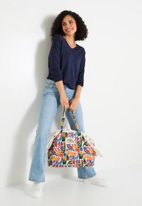 Mujer de pie contra una pared blanca, sonriendo, vestida con un suéter azul marino, jeans de mezclilla claros, zapatillas blancas, sosteniendo un bolso de tote con un estampado floral y un parche de texto.