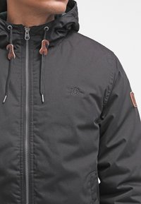 Czarna kurtka na zamek z kapturem, z troczkami zakończonymi brązowymi końcówkami, bocznymi kieszeniami i logo na piersi. Materiał wydaje się gładki.