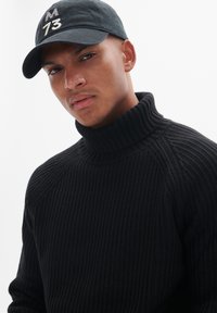 Suéter de punto acanalado negro con cuello alto, llevado debajo de una gorra de béisbol negra con letras bordadas en blanco.