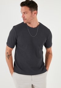 Tricou scurt de culoare gri închis, cu un model texturat în forma de vafă și guler rotund, asortat cu pantaloni de culoare deschisă. Accent minimal cu un lănțișor argintiu.