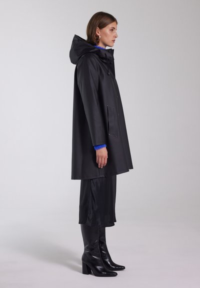 Svart vattentät regnjacka med huva, med en frontknäppning, sidofickor och en slät textur. Bar över mörka kläder och knähöga stövlar.