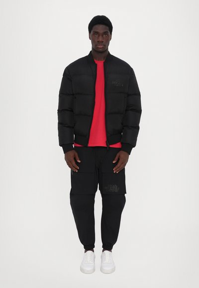 Zwarte pufferjack met geribde manchetten en een rood T-shirt eronder, gecombineerd met zwarte cargo-broeken met zijzakken en witte sneakers.