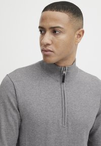 Grauer Zip-Pullover mit hohem Kragen, ausgestattet mit einem Metallreißverschluss und gerippten Bündchen. Weiche Textur, figurnahes Design, minimale Nähdetails.