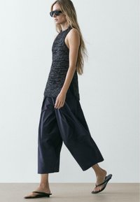 Kvinde går indenfor iført ærmeløs mørk striktop, vidbenede sorte bukser, sorte flip-flops og store mørke solbriller.