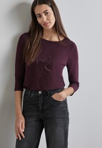 Burgunderfarbener Langarmshirt mit Rundhalsausschnitt und bedrucktem Text, kombiniert mit schwarzen Jeans aus Denim mit Fronttaschen und Knopfverschluss.