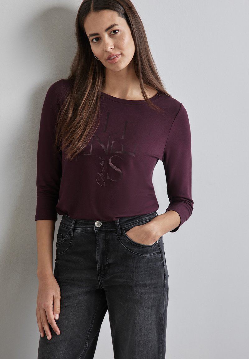 Burgunderfarbener Langarmshirt mit Rundhalsausschnitt und bedrucktem Text, kombiniert mit schwarzen Jeans aus Denim mit Fronttaschen und Knopfverschluss.