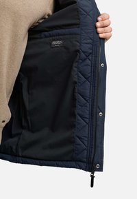 Chaqueta acolchada de color navy con forro interior negro, cierre de cremallera y broches de metal, que presenta una etiqueta cosida en el lado izquierdo.