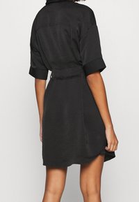 Black dress with short sleeves, a collar, and a wrap tie at the waist. Features a textured fabric and a slightly curved hem.