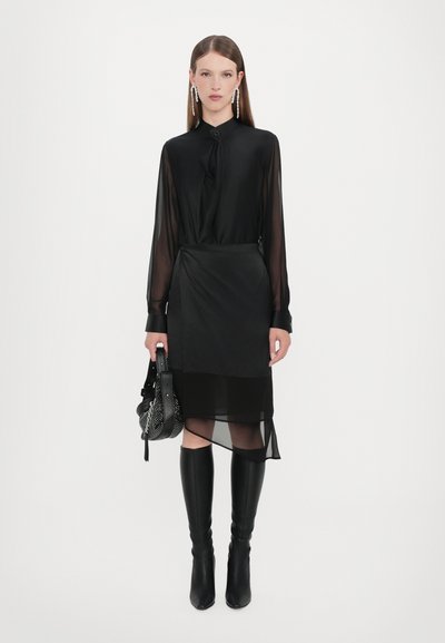 Black sheer blouse with button closure, paired with a layered black skirt. Knee-high black boots. Holding a black textured handbag.