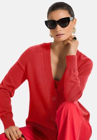 Rode cardigan met een V-hals, geribde manchetten en twee knopen, gecombineerd met een bijpassende top en broek. Zwarte cat-eye zonnebril en statement oorbellen.