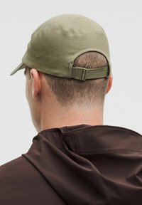 Gorra verde oliva con una tela texturizada, visera curvada y correa ajustable en la parte posterior, combinada con una chaqueta marrón oscuro.